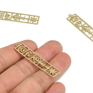 Könnte beinhalten: Goldfarbene rechteckige Charms mit ausgeschnittenen Designs. Die Charms zeigen verschiedene Formen, darunter eine Sonne, ein Blatt und ein Stern. Ein Charm wird in einer Hand gehalten, während zwei weitere in der Nähe verstreut sind. Diese Charms sind ideal für die Schmuckherstellung.