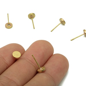 May include: A collection of small, gold-coloured stud earrings with a flat, circular design are displayed against a white backdrop. One earring is held between fingers. The earrings have a post for insertion into the ear. These earrings are suitable for everyday wear.