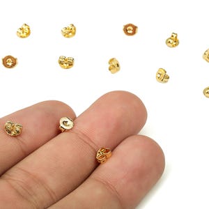 以下が含まれることがあります： 白い背景に、ゴールドカラーのイヤリングキャッチ（バタフライバックとも呼ばれる）のコレクションが散らばっています。 いくつかは焦点が合っており、他はわずかにぼやけています。 イヤリングキャッチの一部は手で持たれています。