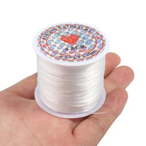 Puede incluir: Un carrete de hilo de cristal transparente, fuerte y elástico. La etiqueta dice "STRONG & STRETCHY" y "CRYSTAL STRING" en inglés y japonés. El carrete se sostiene en una mano.
