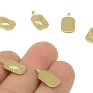 May include: Gold-colored rectangular pendants with a heart design and a small loop at the top. Some pendants are held in a hand. The pendants are made of metal and have a matte finish. The pendants are small, approximately 0.75 inches long.