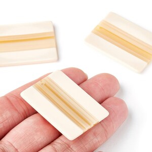 May include: Ivory-coloured rectangular beads with a light brown stripe design. The beads are shown in a hand, with two additional beads in the background. These beads are suitable for jewellery making.