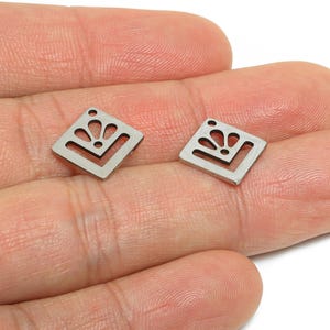 May include: Two small, silver-colored, square-shaped earring charms. Each charm features a decorative cutout design resembling a stylized flower or sunburst within a V-shaped frame. The charms have a small hole at the top for attachment.