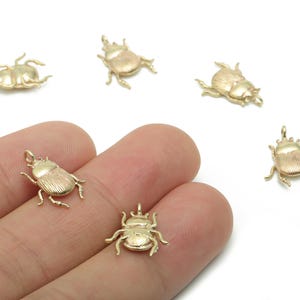 Puede incluir: Amuletos de escarabajo de color dorado. Los amuletos son pequeños, detallados y tienen un lazo en la parte superior para sujetarlos a las joyas. Los escarabajos están en varias poses, con sus patas y antenas visibles. El fondo es blanco.