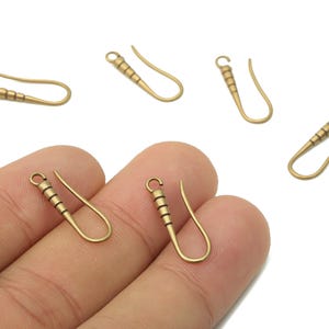 May include: A collection of gold-coloured earring hooks. The hooks feature a curved design with a decorative, ridged section near the top. One pair of hooks is held in a hand, with others scattered around.