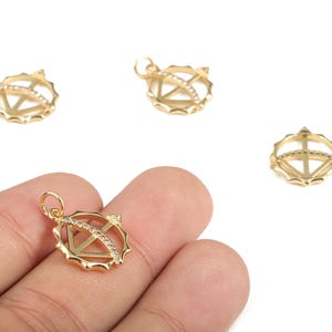 Könnte beinhalten: Goldfarbene Anhänger-Charms mit geometrischem Design. Die Charms haben einen kreisförmigen Rahmen mit rautenförmigem Innenraum und sind mit kleinen, klaren Steinen verziert. Die Charms werden vor weißem Hintergrund gezeigt.