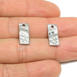 Puede incluir: Dos pequeños dijes rectangulares de metal plateado con textura martillada. Cada dije tiene un pequeño agujero en la parte superior. Los dijes se sostienen en la mano de una persona.
