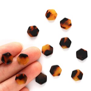 May include: A collection of hexagonal beads in a tortoiseshell pattern. The beads are a mix of amber and dark brown, with a glossy finish. Some beads are held in a hand, while others are scattered on a white surface.