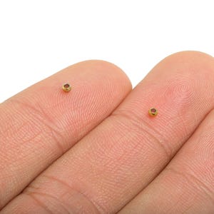 May include: Two tiny, gold-colored metal beads with a reddish-brown center are shown resting on human fingertips. The beads are small, round, and have a polished appearance. The background is plain white, emphasizing the details of the beads.