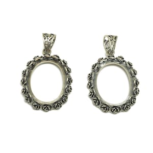 Puede incluir: Dos colgantes ovalados plateados con detalles florales en los bordes. Cada marco tiene una argolla decorativa en la parte superior para sujetarlo a una cadena. Los colgantes se muestran sobre un fondo blanco.