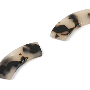 Może przedstawiać: Dwa brązowe i czarne plastikowe spinki do włosów w stylu imitującym skorupę żółwia. Spinki są zakrzywione i mają otwór na górze.