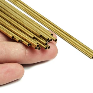 May include: A selection of small, hollow, brass tubes. The tubes are cylindrical, with a golden hue, and are held in a hand against a white backdrop. The tubes are of varying lengths and are grouped together.