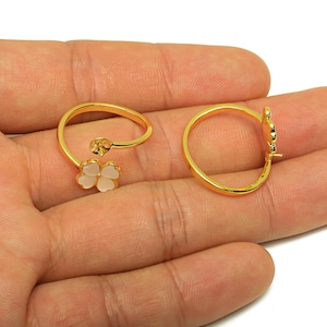 Könnte beinhalten: Zwei goldfarbene Ringe. Einer hat ein vierblättriges Kleeblatt-Design mit weißen Blütenblättern. Der andere Ring ist ein schlichtes Band mit einem kleeblattförmigen Akzent. Beide Ringe werden auf einer Hand gezeigt.