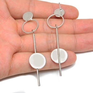 Pode incluir: Um par de brincos pendurados em tons de prata. Cada brinco apresenta um pequeno disco, um aro circular, uma longa barra vertical e um disco maior na parte inferior. Os brincos são exibidos em um fundo claro.