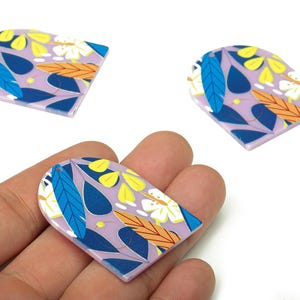 Puede incluir: Tres colgantes decorativos con forma de arco y diseño floral. Los colgantes presentan un fondo lavanda con motivos florales y de hojas azules, amarillos, naranjas y blancos. Un colgante se sostiene en una mano.