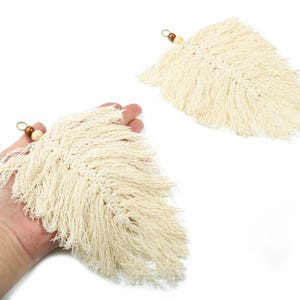 May include: Two cream-colored macrame feather-shaped wall hangings. One is held in a hand, the other is separate. Each has a small wooden bead and a metal loop at the top for hanging. The feathers have a textured, fringed design.