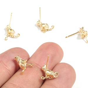 Op de afbeelding: Goudkleurige katvormige oorstekers. De oorbellen hebben een textuuroppervlak en een pin voor het inbrengen. Verschillende oorbellen zijn verspreid op een wit oppervlak, waarvan er twee in een hand worden gehouden.