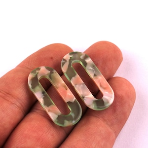 Peut inclure: Deux boucles d'oreilles ovales avec un motif marbré vert, rose et blanc. Les boucles d'oreilles sont tenues dans une main sur fond blanc. Ces boucles d'oreilles conviennent à la fabrication de bijoux.