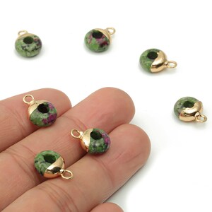 Può includere: Piccole perline rotonde di pietre preziose con un cappuccio e un anello in metallo dorato. Le perline sono verdi con inclusioni rosse e viola. I cappucci dorati coprono una parte della perlina.