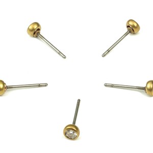 May include: Five pairs of gold stud earrings with clear gemstones. Each earring has a small, round, clear gemstone set in a gold setting. The earrings are on a white background.