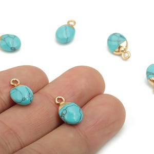 May include: A collection of turquoise-colored, oval-shaped gemstone charms with gold-colored metal caps and loops. The charms are a light blue with dark blue veining. The gold-colored metal loops are attached to the top of each charm.