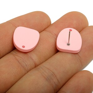 Pode incluir: Dois brincos de botão rosa claro. Um brinco tem formato arredondado com um pequeno ponto vermelho. O outro brinco tem formato semelhante com um pino prateado e um pequeno ponto vermelho. Os brincos são mostrados em uma mão.