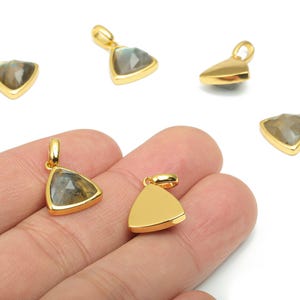 Puede incluir: Colgantes triangulares dorados con un centro de piedras preciosas. Los colgantes tienen un lazo en la parte superior para una cadena. Un colgante se sostiene en una mano. Las piedras preciosas tienen tonos grises, verdes y marrones. Joyas.