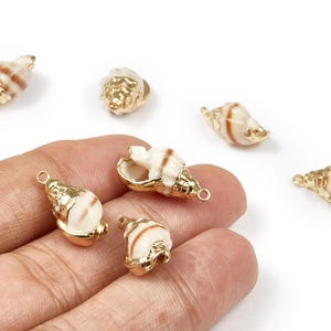 Könnte beinhalten: Mehrere kleine Muschel-Anhänger mit goldfarbenen Akzenten. Die Muscheln haben eine natürliche, cremefarbene Basis mit braunen Streifen. Jeder Anhänger hat eine kleine Schlaufe zum Befestigen an Schmuck oder anderen Bastelarbeiten.