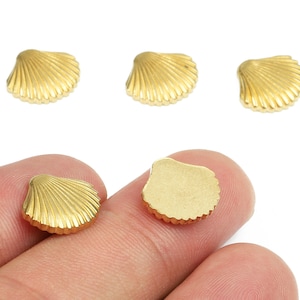 Op de afbeelding: Goudkleurige schelpenbedels. De bedels zijn klein, met een gedetailleerde, geribbelde textuur. Verschillende liggen verspreid op een witte achtergrond, terwijl er twee tussen vingers worden gehouden. Geschikt voor het maken van sieraden of knutselprojecten.