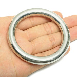 Peut inclure: Un bracelet jonc argenté brillant. Le bracelet circulaire est épais et lisse, avec une finition polie. Le bracelet est tenu dans une main ouverte sur fond blanc. Ce bijou est adapté au port quotidien.