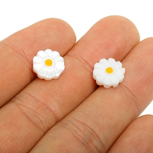 May include: Two small, white daisy-shaped beads with yellow centres are displayed. The beads are held in a person's hand, showcasing their delicate floral design. These beads are suitable for jewellery making or craft projects.