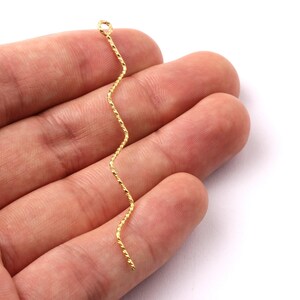 May include: A gold-colored, wavy jewelry finding with a textured surface. The finding has a small loop at the top and a zig-zag design. The jewelry finding is held in a hand against a white background.