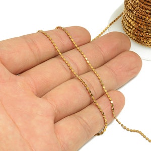 May include: A gold-colored chain with small, round links. The chain is lying on a hand, and a spool of the chain is visible in the background.