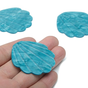 May include: Three turquoise seashell-shaped craft beads. The beads have a scalloped edge and a textured, marbled appearance. One bead is held in a hand. These beads are suitable for jewelry making and other craft projects.