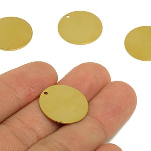 May include: Close-up of several gold-coloured, round metal charms with a small hole at the top for stringing. One charm is held in a hand. These blank pendants are ideal for jewellery making and crafting.