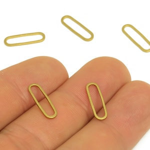 May include: Several small, gold-colored oval-shaped jewelry findings are displayed against a white background. Two of the findings are held in a person's hand. The findings are open ovals, perfect for jewelry making and crafting.