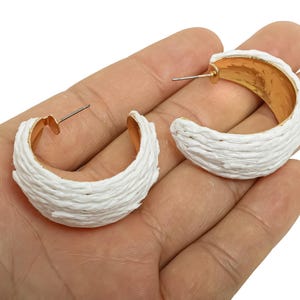 May include: A pair of white hoop earrings with a textured, woven appearance. The inner portion of the earrings is gold-coloured, and the posts are silver. The earrings are displayed on a hand.