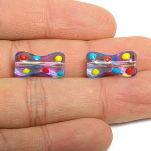 May include: Two clear, bow-tie shaped beads with colorful polka dots in red, yellow, and blue. The beads are held in a person's hand, showcasing their iridescent quality and design. These beads are ideal for jewelry making.