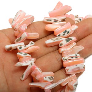 May include: A coral necklace featuring irregular, oblong beads in shades of pink, white, and gray. The beads are strung together, creating a unique and natural jewelry piece. The necklace is shown against a neutral background.