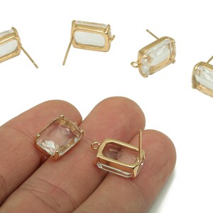 May include: Gold-tone rectangular stud earrings with clear, faceted crystal inserts. The earrings have a post backing and a small loop for attaching charms. Several earrings are displayed, with two held in a hand.