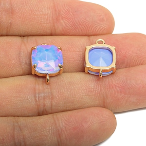 Könnte beinhalten: Zwei quadratische, irisierende blau-rosa Edelsteinanhänger mit goldfarbenen Metalleinfassungen. Jeder Anhänger hat oben und unten eine Schlaufe zum Befestigen an einer Kette oder einem anderen Schmuckbestandteil. Die Edelsteine sind facettiert.