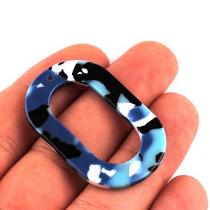 Peut inclure: Un pendentif ovale à motifs bleus, noirs et blancs. Le pendentif est tenu dans une main et présente un motif marbré. La palette de couleurs comprend des nuances de bleu, de noir et de blanc. Le pendentif a un trou en haut.