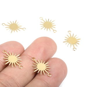 Könnte beinhalten: Goldfarbene Sonnenstrahl-Charms mit Verbindungsschlaufen. Die Charms haben ein sonnenähnliches Design mit spitzen Strahlen, die sich von einem zentralen Kreis aus erstrecken. Mehrere Charms sind auf weißem Hintergrund verstreut.