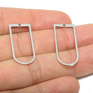 Puede incluir: Dos colgantes plateados de marco abierto. Cada colgante tiene una parte superior rectangular con un pequeño agujero y una parte inferior redondeada. Los colgantes se muestran sobre un fondo neutro.