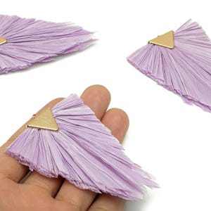 May include: Three lavender-colored raffia fan-shaped earring components with gold-tone triangle connectors. The earrings are displayed on a white background. These components are suitable for jewelry making.