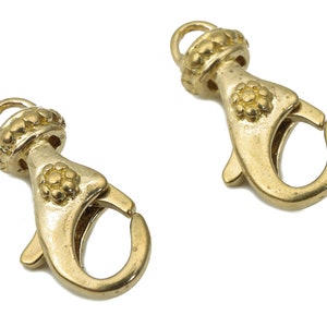 May include: Two gold-tone metal lobster clasps with decorative accents. Each clasp has a round, textured ring and a flower-shaped design.