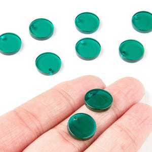 Könnte beinhalten: Eine Sammlung von smaragdgrünen, kreisförmigen Acryl-Anhängern mit einem kleinen Loch oben. Mehrere Anhänger sind auf einer weißen Oberfläche verstreut, während zwei in einer Hand gehalten werden. Ideal für Schmuckherstellung und Basteln.