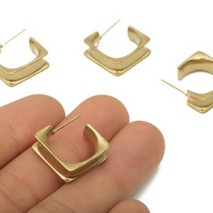 Könnte beinhalten: Goldfarbene quadratische Creolen. Die Ohrringe haben ein geschichtetes Design mit quadratischer Form und einem Stecker zum Durchstechen. Die Ohrringe werden vor weißem Hintergrund gezeigt. Die Ohrringe haben ein schlichtes, elegantes Design.