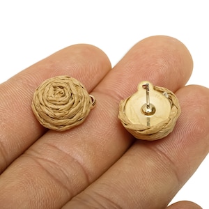 May include: A pair of round stud earrings. Each earring has a woven, beige design, similar to a rose. The earrings have a gold-coloured backing and post. The earrings are shown on a hand.