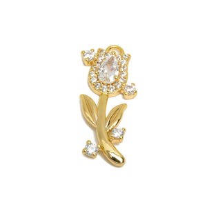 May include: A gold-colored floral charm with a tulip-like design. The flower head is encrusted with clear crystals, and the stem features two leaves and additional crystals. The charm is set against a white background.